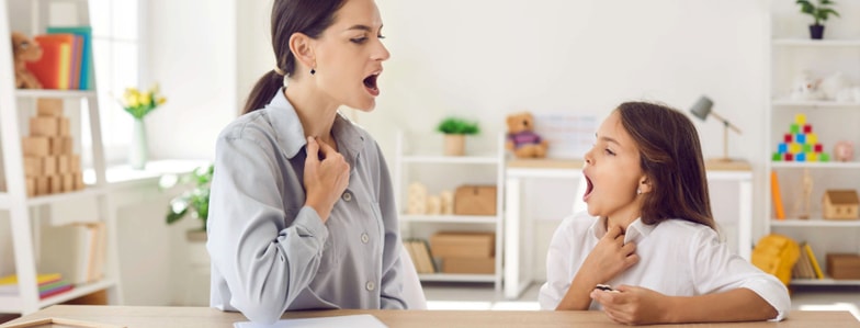 A woman and a girl touch their throats and vocalize together at a desk in a bright, tidy medbridge therapy room.