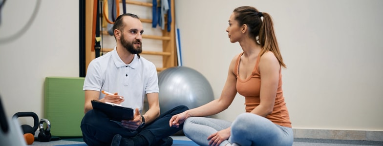 A man with a clipboard talks to a woman in athletic wear on a gym floor, with medbridge digital healthcare and exercise equipment visible.