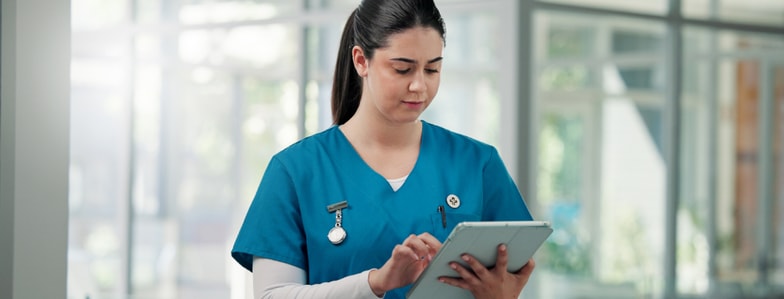 A nurse in blue scrubs uses a tablet with medbridge software in a bright healthcare facility.