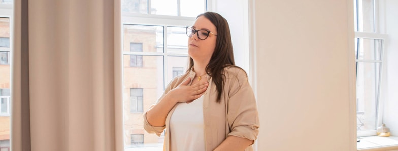 A woman with glasses stands indoors by a window, eyes closed and hand on chest, looking calm and thoughtful—reflecting the mindful approach promoted by medbridge.