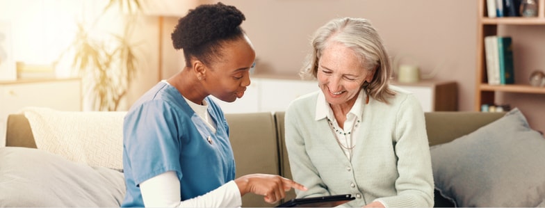 A healthcare worker in blue scrubs shares a tablet with an older woman, both smiling as they use medbridge tools.