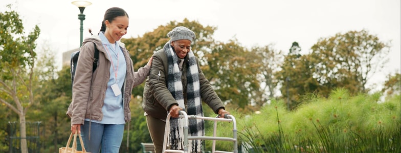 A healthcare worker helps an older woman with a walker in a park.