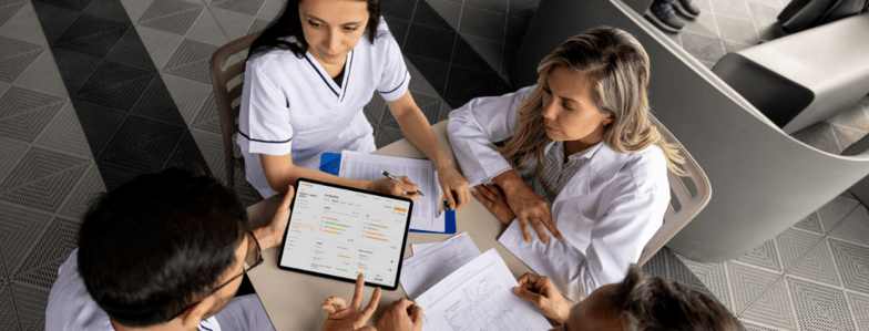 Four people in white uniforms sit at a table, one holding a tablet showing medbridge patient care software.