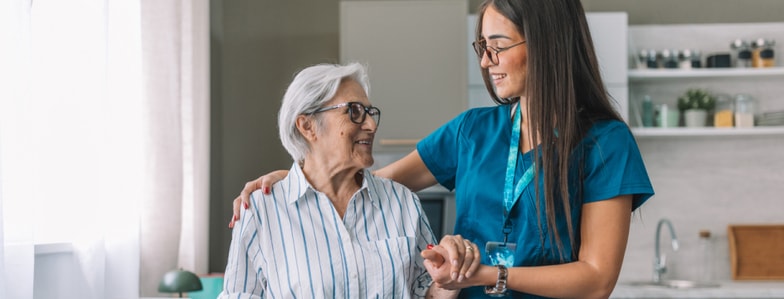 A caregiver in blue scrubs smiles while using Medbridge software to hold an elderly woman’s hand.